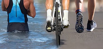 Das Bild zeigt Ausschnitte von Leuten, welche die drei Sportarten die zu Triathlon gehören ausüben. Vonlinks nach rechts sieht man: Den Rücken eine Person welche schwimmt, die Füße einer Person, welche Rad fährt, auf den Pedalen, die Füße einer Person die läuft