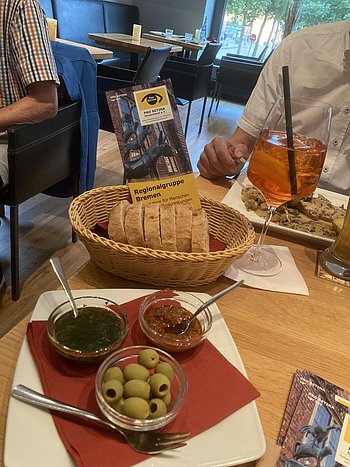 Ein Teller mit Pesto und Oliven, dahinter steht ein Brotkorb mit rot. Darin steckt ein Flyer der Regionalgruppe Bremen.