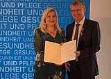 Melanie HUmkl neben Franz BAdura - Beide zeigen die Verleihungsurkunde für das Bundesverdienstkreuz in die Kamera.