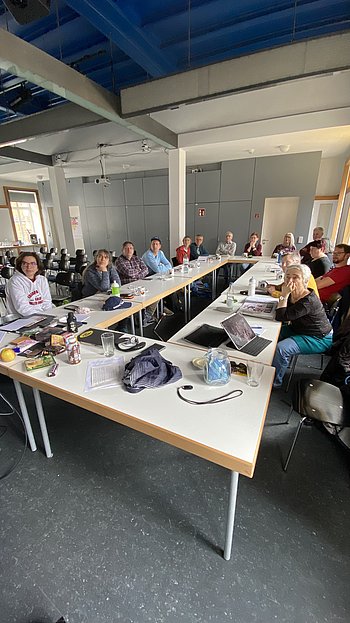 Die Teilnehmenden sitzen in U-Form an Tischen und schauen nach vorne