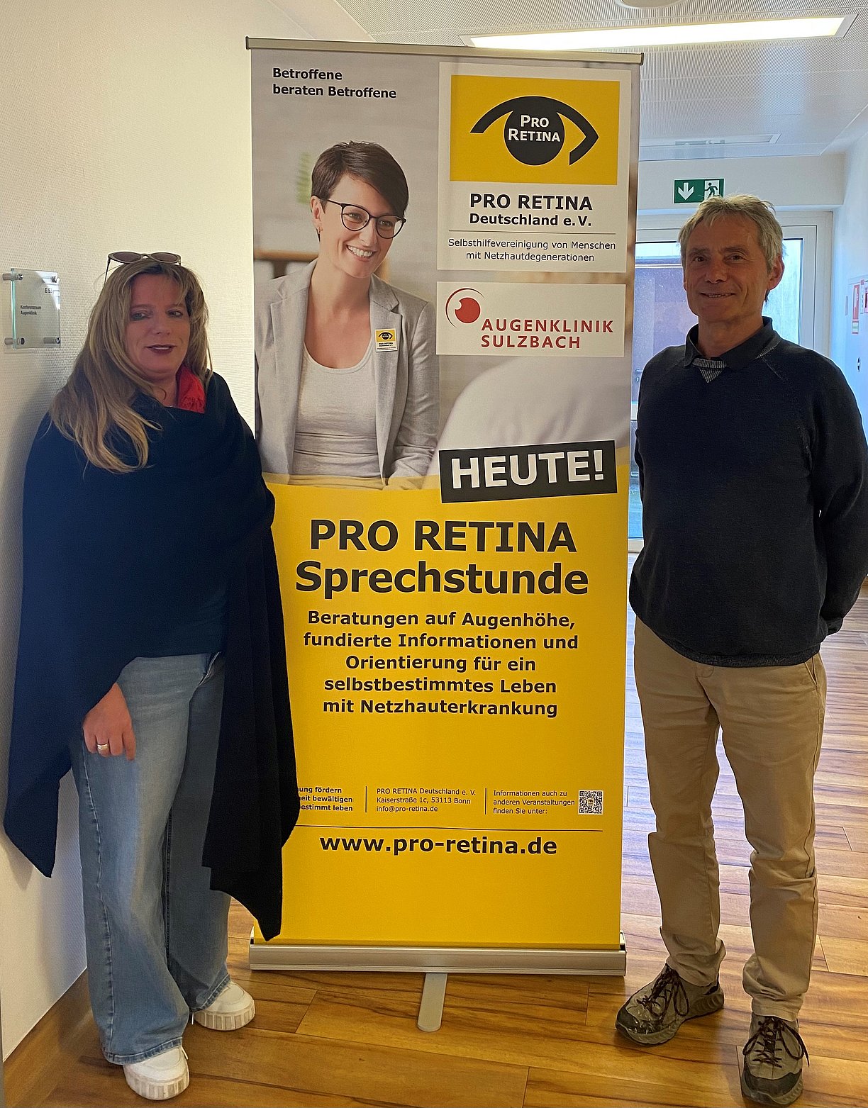 Marion Palm Stalp und Martin Haupenthal stehen neben einem PRO RETINA Aufsteller in der Augenklinik Sulzbach
