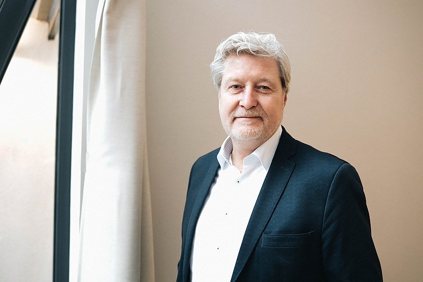 Auf dem Bild zu sehen ist Thomas Reichel, ein Mann mittleren Alters. Seine Haare sind weiß und er hat einen leichten, gepflegten Bart. Er trägt ein Jackett und ein Hemd.