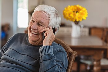 Ein älterer Mann sitzt zu Hause und telefoniert lächelnd mit einem Smartphone.
