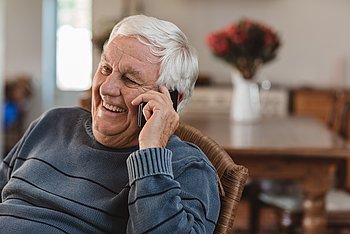 Ein älterer Mann sitzt zu Hause und telefoniert lächelnd mit einem Smartphone.