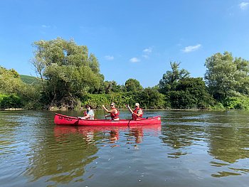 Zwei Personen paddeln in einem roten Kanu auf einem ruhigen Fluss. Beide tragen Schwimmwesten, das Wetter ist sonnig, und am Ufer sind grüne Bäume und Sträucher zu sehen.