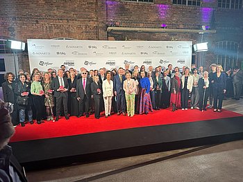 Auf dem Abschluss-Pressebild der 23. Hörfilmpreisverleihung stehen die Gastgeber, die Moderatorin, die Laudatoren, Jurymitglieder und Preisträger auf dem roten Teppich vor der Pressewand im Scheinwerferlicht. Die Namen der Sponsoren, darunter die Aktion Mensch und Pfizer, sind auf der Wand zu lesen.  