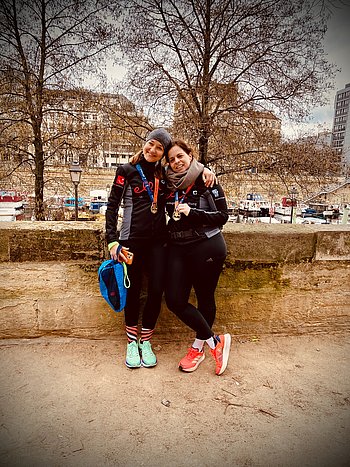 Zwei junge Frauen mit Laufkleidung stehen lächelnd Arm in Arm nebeneinander. Sie tragen Medaillen um den Hals. 