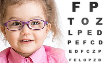 Smiling girl putting on glasses with blurry eye chart behind