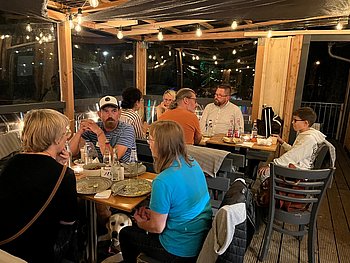 Am Freitagabend gab gab es ein leckeres Grillbuffet im Bootshaus des Ufercafes. Teilnehmer sitzen an Tischen und unterhalten sich. Draußen ist es dunkel, innen ist eine Lichterkette an.