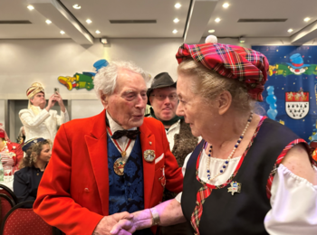 Foto: Links auf dem Bild ist Ludwig Sebus, ein älterer Mann mit weißem Haar. Er trägt ein rotes Jacket, an welches ein Karnevalsorden angesteckt ist und einen weiteren Orden um den Hals. Gerade schüttelt er die Hand von Ute Palm (links), welche ebenfalls einen Orden angesteckt hat und ein Kostüm mit einem karierten Hut trägt