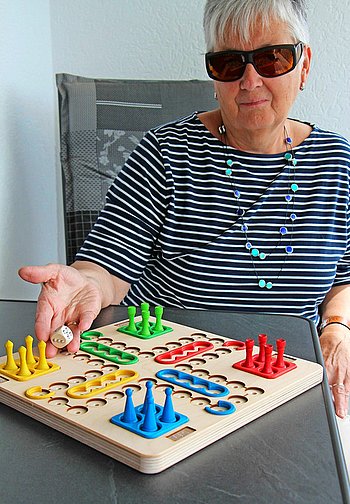 Foto von Brigitte Bettscheider zeigt Roswitha Karst an einem Mensch-ärgere-dich-nicht-Spiel, das speziell auf die Bedürfnisse von Sehbehinderten und Blinden ausgerichtet ist.<﷯