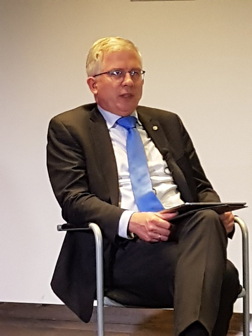 Mann mit weißen Haare und Brille im dunklen Anzug, helles Hemd, blaue Krawatte, sitzt auf einem Stuhl und hat eine Mappe auf dem Schoß.