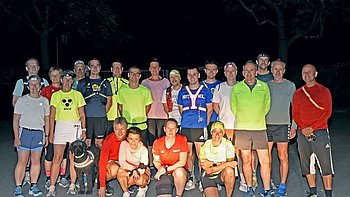 Gruppenfoto von 23 Läuferinnen und Läufern beim Moonlight-Run in Soest. Die Teilnehmenden stehen dicht nebeneinander auf einem dunklen Parkplatz, einige tragen helle oder reflektierende Sportkleidung und Stirnlampen. Vorne sitzt ein schwarzer Blindenführhund mit reflektierendem Geschirr. Alle lächeln in die Kamera. Das Foto wurde bei Nacht aufgenommen und zeigt eine fröhliche, inklusive Sportgemeinschaft.