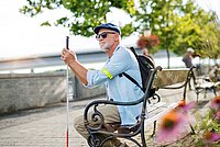 Senior blind man with white cane sitting on bench in park in city.