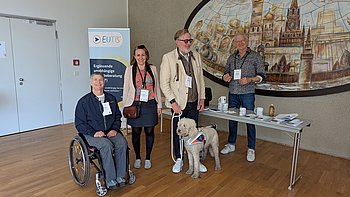 Claudia Posch (EUTB, im Rollstuhl), Zehra Wellmann-Sam (politische Referentin der PRO RETINA), Jörg-Michael Sachse-Schüler (Finanzvorstand der PRO RETINA mit Assistenzhund) und Albrecht Schäufele (EUTB) stehen vor dem EUTB-Banner und Stand im Cafe Moskau in Berlin. Hinten an der Wand ist ein oval eingefasstes, klassisches Gemälde von Moskau zu sehen.