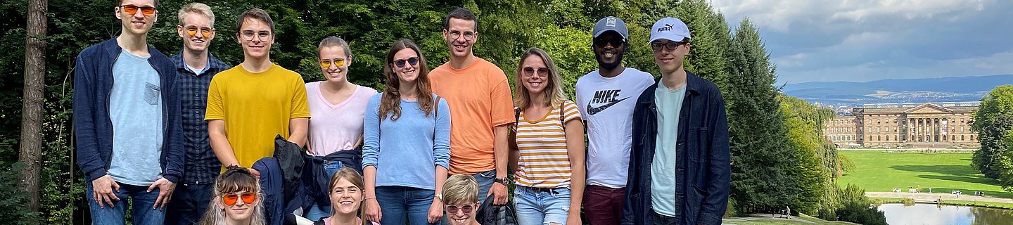 Eine Gruppe junger Menschen, teilweise mit Kantenfilterbrille und Blindenlangstock steht bei gutem Wetter in einem Park. Im Hintergrund sieht man ein Schloss. 