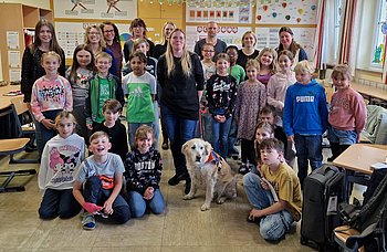 Das Foto zeigt die Schülerinnen und Schüler der Klasse 3c gemeinsam mit ihren Gästen Mandy Kugel, ihrer Begleitperson Doreen Rottmann und der Blindenführhündin Nella. Die Gruppe hat sich im Klassenzimmer für ein Erinnerungsfoto aufgestellt. Die Kinder wirken fröhlich und interessiert. In der Mitte des Bildes sitzt Nella, die ausgebildete Blindenführhündin, aufmerksam auf dem Boden. Das Bild spiegelt die positive und offene Atmosphäre während des Projekttages wider.