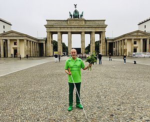 Ein Mann in einem grünen T-Shirt, einer grünen Hose und grünen Schuhen steht vor dem Brandenburger Tor in Berlin. In der rechten Hand hält er einen Langstock, in der linken einen Blumenstrauß.