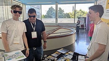 PRO RETINA Mitglied Leander Kronauer klärt zwei junge Interessierte Teilnehmer am Stand über die seltenen degenerativen Netzhauterkrankungen auf. Beide tragen Simulationsbrillen. 