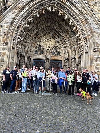 Eine Gruppe von etwa 20 Personen posiert vor dem Eingang einer historischen Kirche. Die Teilnehmer tragen Freizeitkleidung und halten weiße Langstöcke für Blinde. Ein Hund mit einer gelben Weste ist ebenfalls dabei.