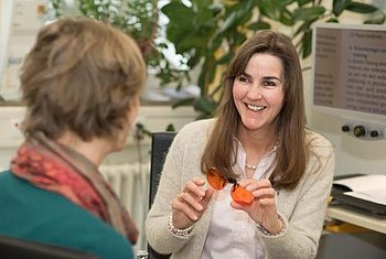 PRO RETINA-Sprechstunde Bonn ©PRO RETINA Eine Frau mit braunen langen Haare sitzt lächelnd vor einer zweiten Frau und zeigt ihr eine Kantenfilterbrille.