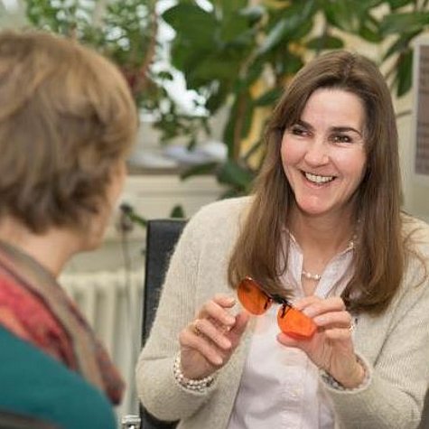 Eine Frau mit braunen langen Haare sitzt lächelnd vor einer zweiten Frau und zeigt ihr eine Kantenfilterbrille. 