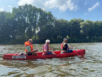 Drei Personen sitzen in einem roten Kanu auf einem Fluss. Zwei von ihnen paddeln, die dritte Person sitzt in der Mitte. Alle tragen Schwimmwesten, das Wetter ist sonnig, und im Hintergrund sind Bäume am Ufer zu sehen.