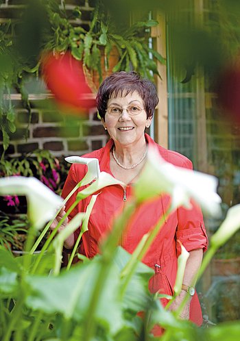 Portrait von Frau Metzner, mit roter Bluse, hinter ein paar dekorativen Blumen stehend. 