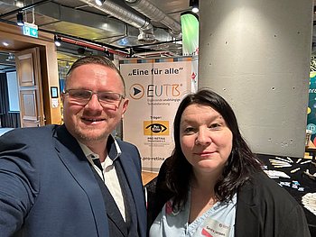 Sylvester Sachse-Schüler und Sandra Jansen stehen vor dem Stand der PRO RETINA. Sylvester trägt ein weißes Hemd mit blauem Sacko. Sandra trägt eine blaue weiß gestreifte Bluse und einen schwarzen Blazer.