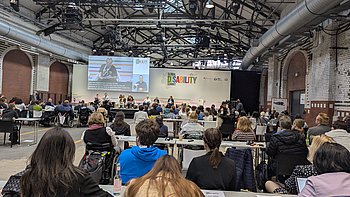 Bild des Panels, in dem darüber diskutiert wird, wie Selbstvertreter erfolgreich Politik beeinflussen können. Die internationalen Panelteilnehmer sitzen vorne am Podium, während der Sprecher auf einer großen Leinwand projiziert wird. Auf der rechten Seite ist das Logo des Disability Summit zu sehen. Die Teilnehmer des Panels sind von hinten in Reihen angeordnet.