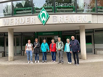 Die Gruppe steht vor dem Weserstadion. Über ihnen ist der Schriftzug Werder Bremen mit der typischen Werder Raute zu sehen