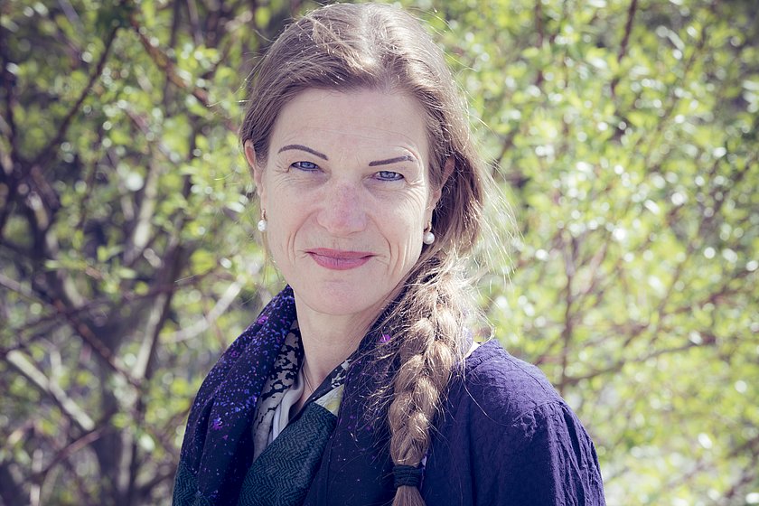 Auf dem Bild zu sehen ist Nicola Janecke, eine Dame mittleren Alters. Ihre blonden Haare sind zu einem Zopf geflochten und liegen über ihrer Schulter. Sie trägt eine dunkle, lilafarbene Bluse und einen Schal in der selben Darbe, außerdem Perlenohrringe. Nicola lächelt freundlich in die Kamera und scheint sich im Freien zu befinden.