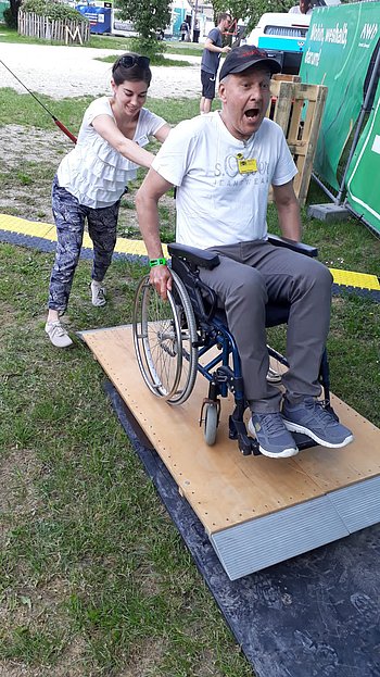 Ein Mann probiert das Rollstuhlfahren und wird von einer Assistentin auf eine Rampe geschoben