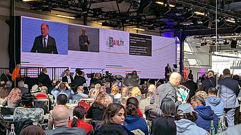 Wir sehen die Bühne der Eröffnungsrede, dahinter eine große Leinwand, so lang wie die Bühne, mit mehreren Screens: Ganz links sieht man den vergrößerten Sprecher, Bundeskanzler Olaf Scholz, rechts daneben den Screen mit dem Gebärdendolmetscher, noch weiter rechts das Logo des Global Disability Summit und ganz rechts die Schriftübersetzung. Vorne steht der Redner am Pult. Im vorderen Teil des Bildes sieht man die Sitzreihen und das Publikum von hinten. Der postindustrielle Raum ist lichtdurchflutet. 