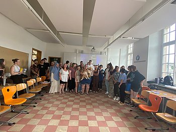 Gruppenfoto von etwa 25 Personen in einem Klassenraum mit großen Fenstern, einige sitzen auf orangen Stühlen, eine Person hält einen Hund
