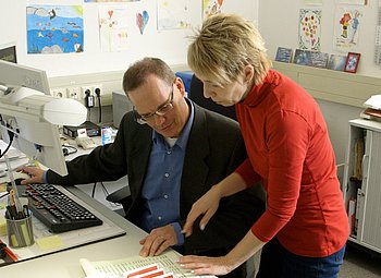 Ein Mann mit schwarzem Jacket und Brille sitzt an einem Schreibtisch mit Computer. Neben ihm steht eine Frau mit roter Bluse und schaut mit ihm gemeinsam auf ein Dokument.