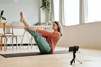 Eine Frau in SPortkleidung sitzt auf einer Yogamatte. Sie hat die Beine schräg nach oben gestreckt, die Arme parallel zum Boden gehoben.