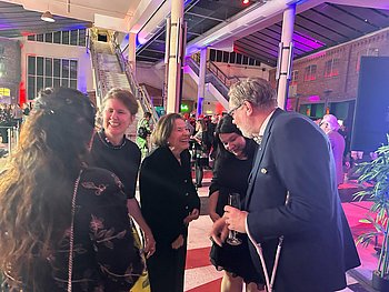 Die Vertreter von PRO RETINA Deutschland e.V. sind auf der Aftershow-Party der Hörfilmpreisverleihung im Gespräch mit Eva Luise und Dr. Ulrike Köhler. Das hohe Foyer ist stimmungsvoll in blaues und rotes Licht getaucht, die Gäste sind in eleganter Abendgarderobe, die Damen in Kleidern und die Herren im Anzug mit Fliege. Auf dem Bild sind von links nach rechts zu sehen: Zehra Wellmann-Sam, Dr. Ulrike Köhler, Eva Luise Köhler, Dr. Sandra Jansen, Jörg-Michael Sachse-Schüler mit Assistenzhund.