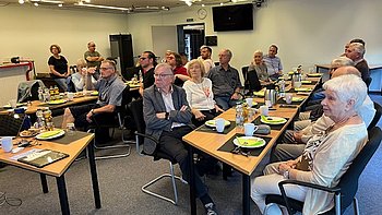 Konferenzraum mit mehreren  Menschen, die an Tischen mit Kaffee und Gebäck sitzen und aufmerksam nach vorne schauen.