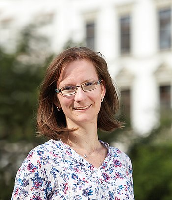 Portrait einer lächelnden, jungen Frau mit braunem, schulterlangen Haar, Brille und einer geblümten Bluse.