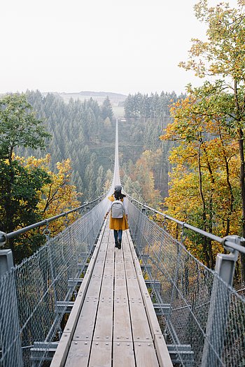 Eine junge Frau sieht man von hinten über eine lange Hängebrücke wandern, sie trägt dabei Wanderkleidung. Die Landschaft im Hintergrund sind herbstgelbe Bäume.