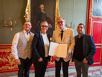 Verleihung des Bundesverdienstkreuzes an Jörg-Michael Sachse-Schüler Gruppenbild von drei Männern und einer Frau in festlicher Kleidung. Die Gruppe steht in einem Saal mit goldenem Stuck und einem Gemälde im Hintergund. Einer der Männer hält eine Urkunde in den Händen. Am rechten Revers trägt er ein rot-goldenes Kreuz.