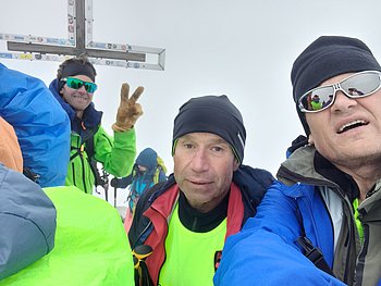 Jubel am Gipfelkreuz. Im Hintergrund steht ein Bergführer und zeigt mit seinen Fingern das Viktoryzeichen. Ein V.