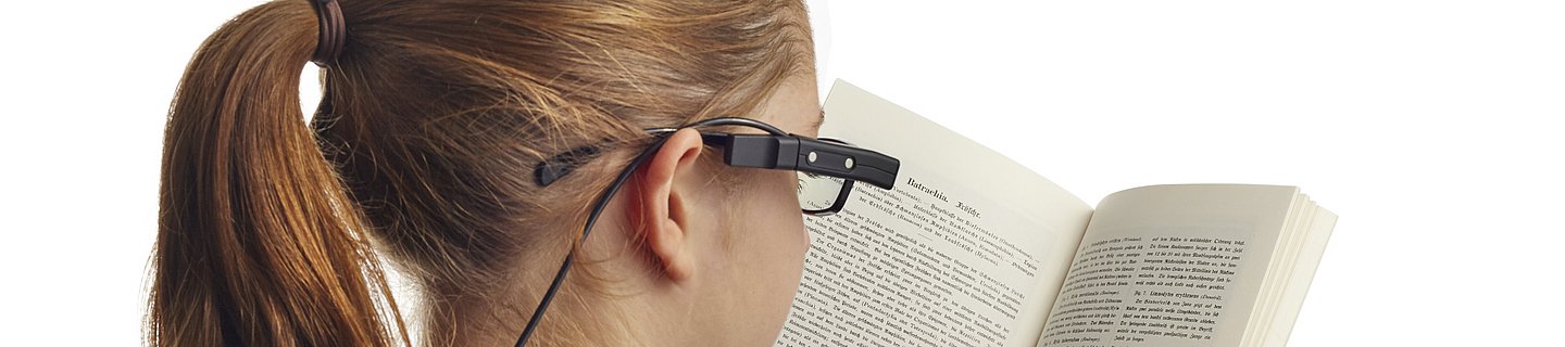 Eine Frau mit blondem Pferdeschwanz und Brille hält ein aufgeschlagenes Buch vor sich und zeigt auf eine Zeile. An ihrer Brille ist eine kleine Kamera befestigt. 