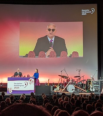 Von links nach rechts: DBSV-Präsident Hans Werner Lange und Moderatorin Nadine Heidenreich stehen in edler Abendgarderobe am Rednerpult, welches sich auf der Bühne befindet. Weiter rechts auf der Bühne stehen die nicht besetzten Musikinstrumente und Musikanlagen der Band Pudeldame. DBSV-Präsident Hans Werner Lange steht mittig am Rednerpult. Er hat ein Mikrofon in der Hand und hält seine  Eröffnungsrede. Moderatorin Nadine Heidenreich steht mit einigem Abstand an der Ecke des Rednerpults und hört aufmerksam zu. Auf einem Teil der großen Leinwand, die sich im hinteren Bereich der Bühne befindet, wird die Rede als Projektion im Großformat übertragen. Im unteren Bildbereich sind zahlreiche Sitzreihen mit Publikum zu sehen. 