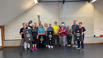 Die Teilnehmenden des Selbstverteidigungskurses der PRO RETINA Regionalgruppe Lörrach/Südschwarzwald stehen gemeinsam im Übungsraum und lächeln in die Kamera. Einige halten Trainingspolster in den Händen.