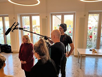 Ein Filmteam mit Kamera, Tonangel und Mikrofon begleitet eine Frau während Dreharbeiten in einem hellen Raum mit großen Fenstern.