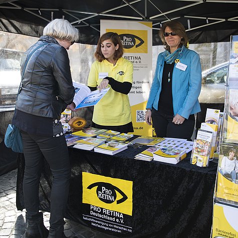 Zwei Frauen beraten eine Frau an einem Infostand mit Broschüren. 