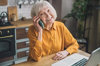 Bild: Eine ältere Frau sitzt telefonierend vor einem Laptop und lächelt in die Kamera.