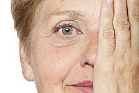 Portrait einer Frau mit kurzem blondem Haar, die sich mit einer Hand das linke Auge zuhält und in die Kamera lächelt.